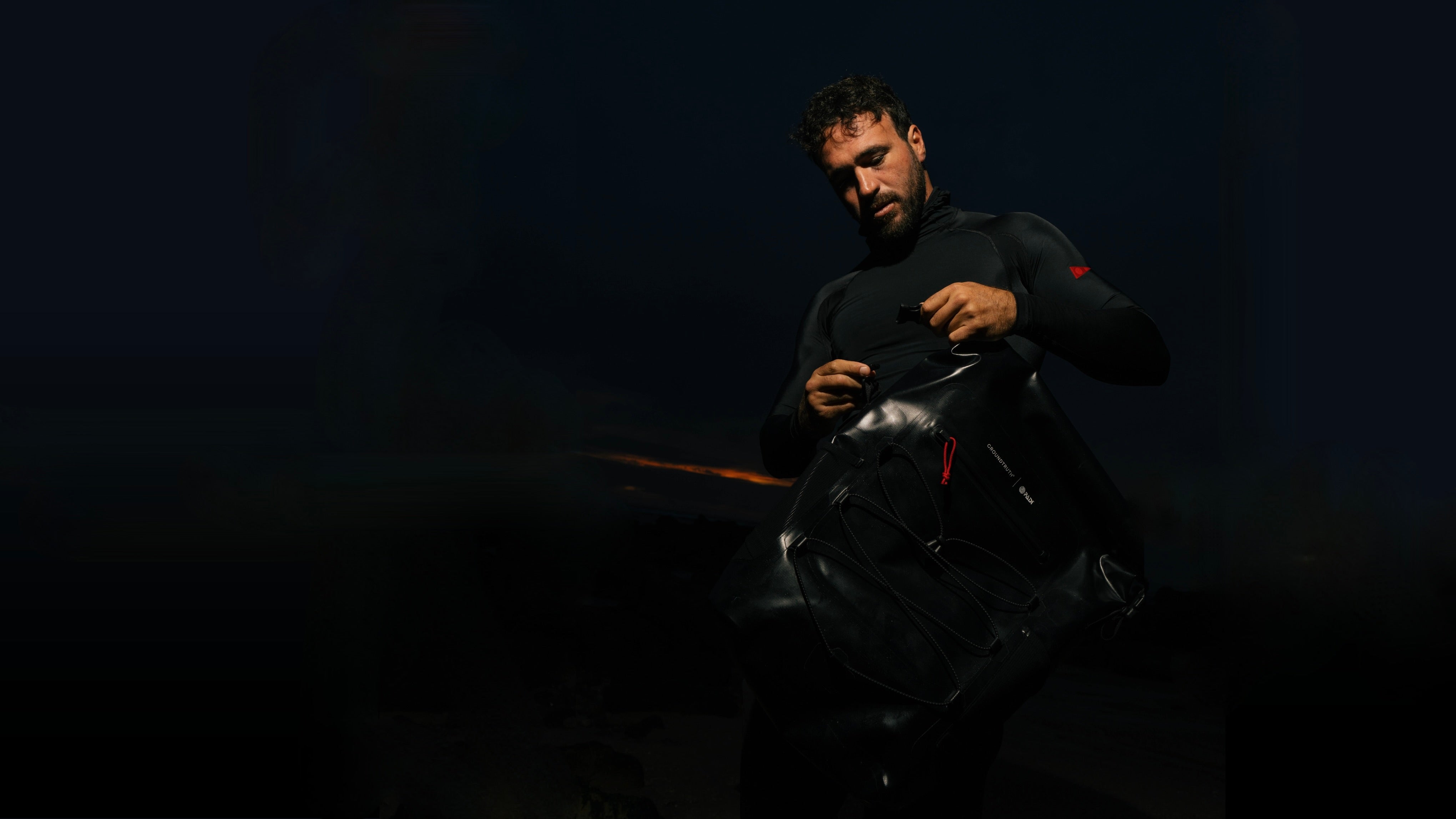 A man in a black long-sleeve top and black pants is shown in very low light, holding and opening a black backpack. A small red logo is visible on the upper sleeve.
