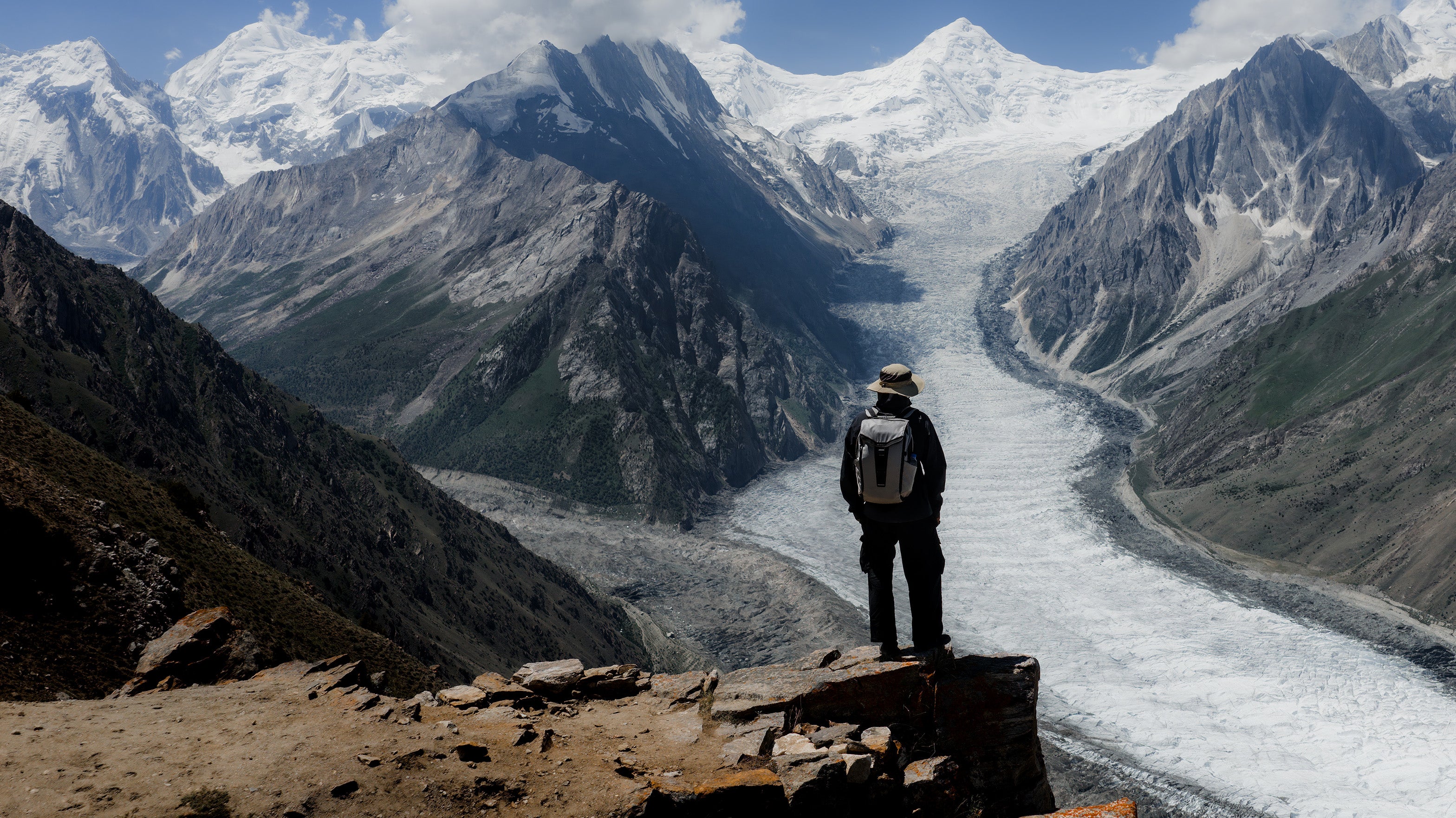 he RIKR 24L Ultimate Backpack being tested with a Person standing on a rocky outcrop overlooking a glacier and mountains.